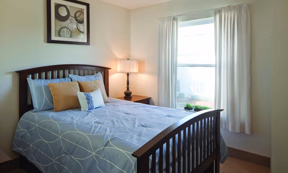 A bright, neatly made bedroom with a wooden bed, blue patterned bedding, nightstand with a lamp, framed artwork, and a window with white curtains.