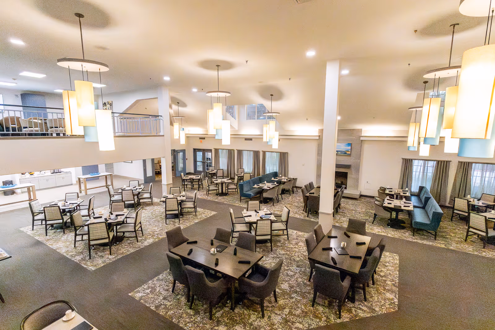 Spacious dining area with multiple tables and chairs arranged neatly on patterned carpet. The room features modern hanging pendant lights, large windows with curtains, and a fireplace in the background. There is an upper level visible with railings and additional seating.