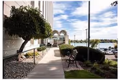 Outdoor walkway alongside a building with a tree and bushes on the left, benches on the right, and a view of a lake with boats in the background under a partly cloudy sky.