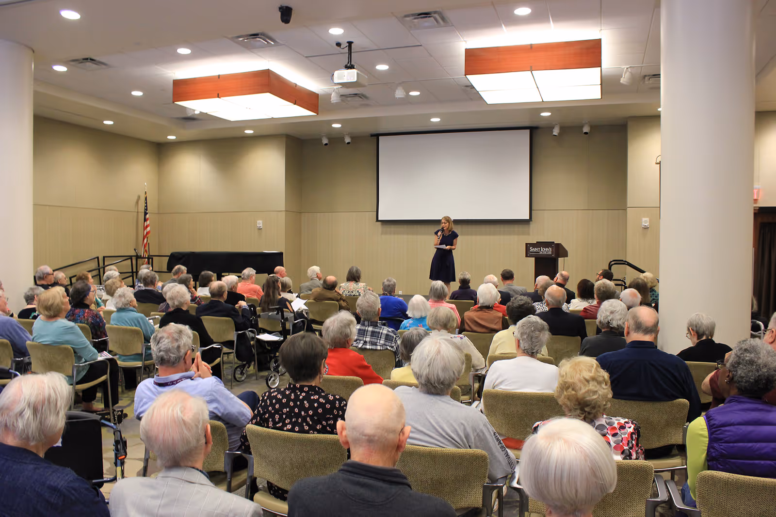 A woman stands on a stage speaking to an audience of elderly people seated in rows of chairs in a large conference room. There is a large projection screen behind her and an American flag to the side. The room is well-lit with ceiling lights and has beige walls.