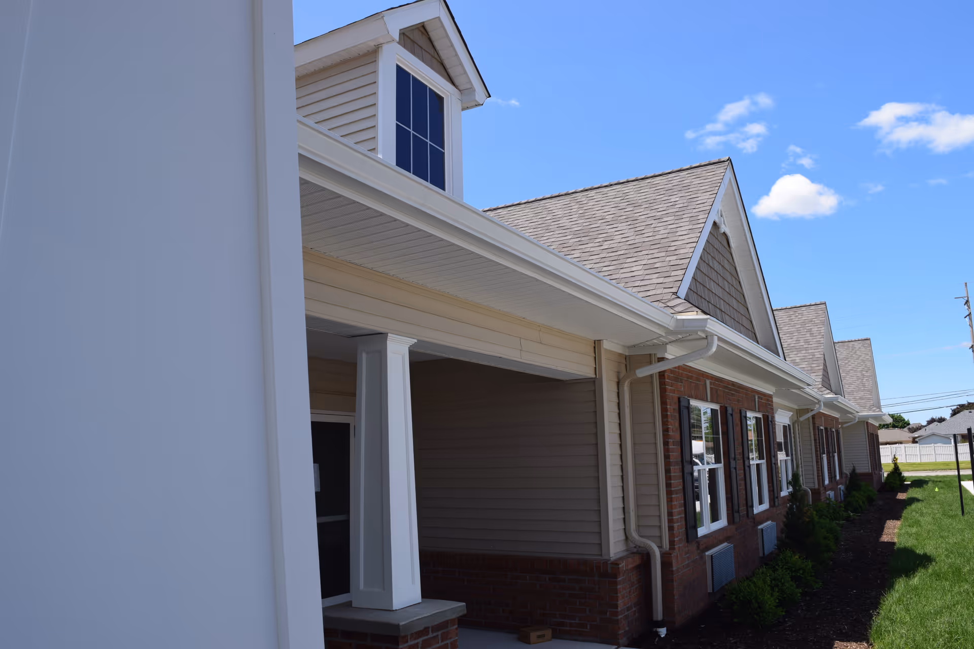 Front exterior of a single-story assisted living building with a covered porch, white columns, brick lower facade, and gabled roof under a blue sky.