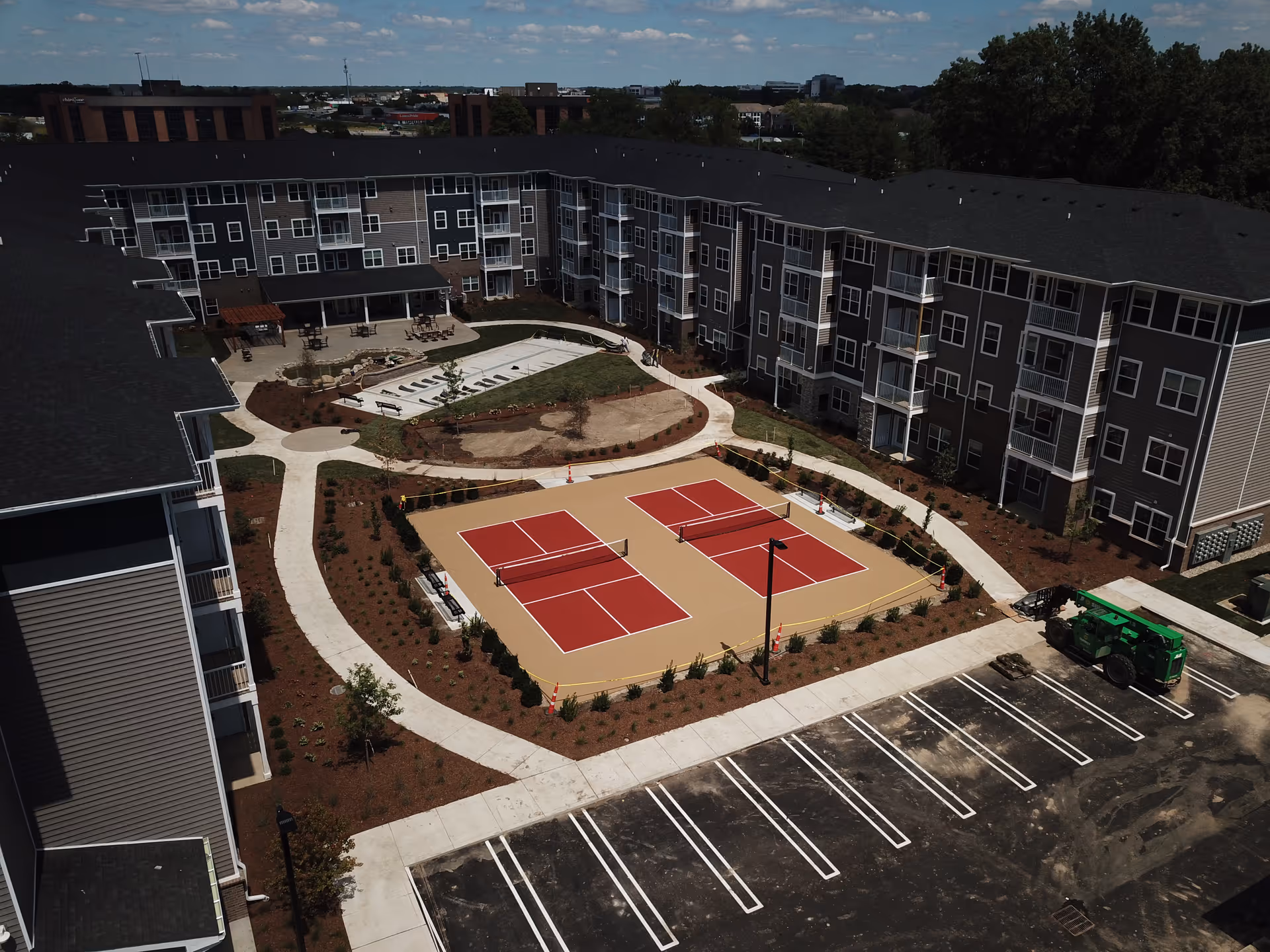 Aerial view of Encore 55+ Boutique Apartments in Plainfield showing a large U-shaped residential building surrounding an outdoor courtyard. The courtyard features two red pickleball courts, walking paths, landscaped areas, benches, and a small patio with tables and chairs. A parking lot is visible in the foreground with a green construction vehicle parked in one of the spaces. Trees and other buildings are visible in the background under a partly cloudy sky.