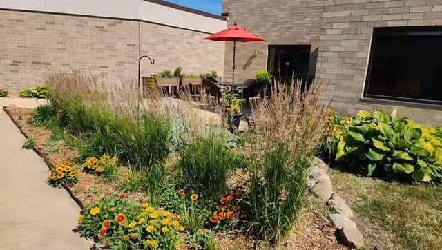 Outdoor garden area with various green plants and colorful flowers along a curved pathway. In the background, there is a patio with a table, chairs, and a red umbrella next to a brick building under a clear blue sky.