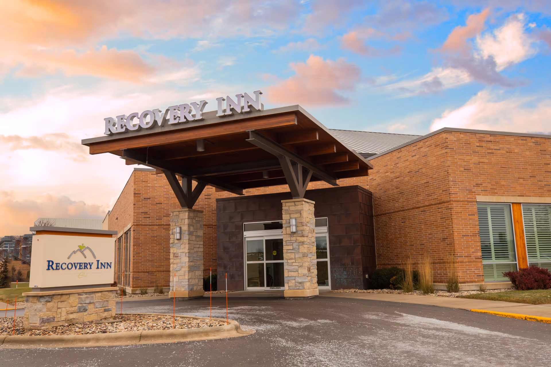 Exterior view of the Recovery Inn building with a covered entrance supported by stone pillars. The building is made of brick and has large windows with shutters. A sign with the Recovery Inn logo is visible near the driveway. The sky is partly cloudy with a warm sunset glow.