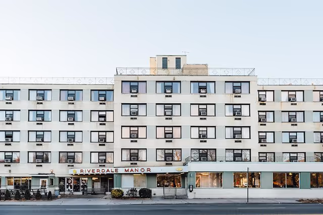 Front exterior view of a multi-story senior living facility named Riverdale Manor with numerous windows, some with air conditioning units, and a clear sky above.