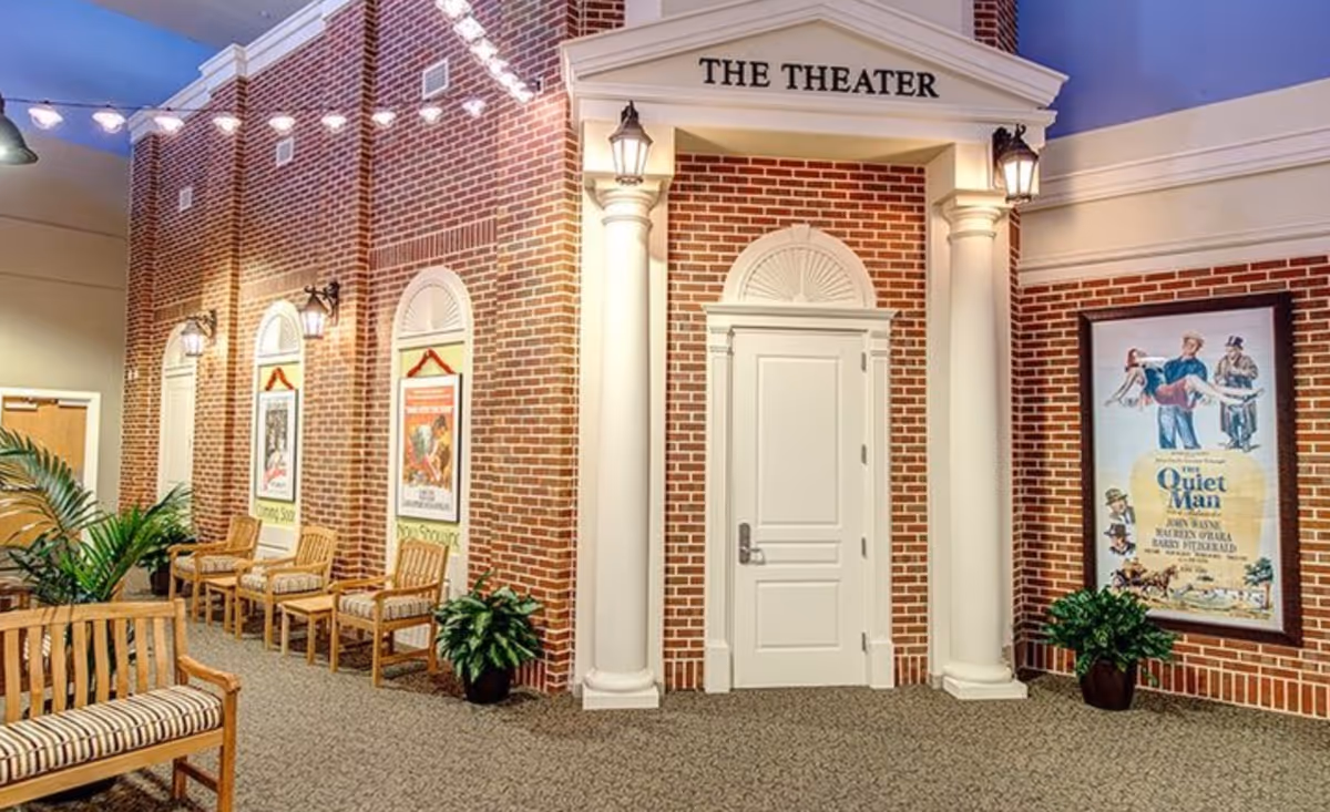 Interior view of a theater entrance in a senior living facility with a white door framed by two large white columns and a brick wall. Above the door is a sign that reads 'THE THEATER'. There are several wooden chairs with cushions along the wall, potted plants, and framed movie posters on the brick walls. String lights hang from the ceiling, creating a warm and inviting atmosphere.