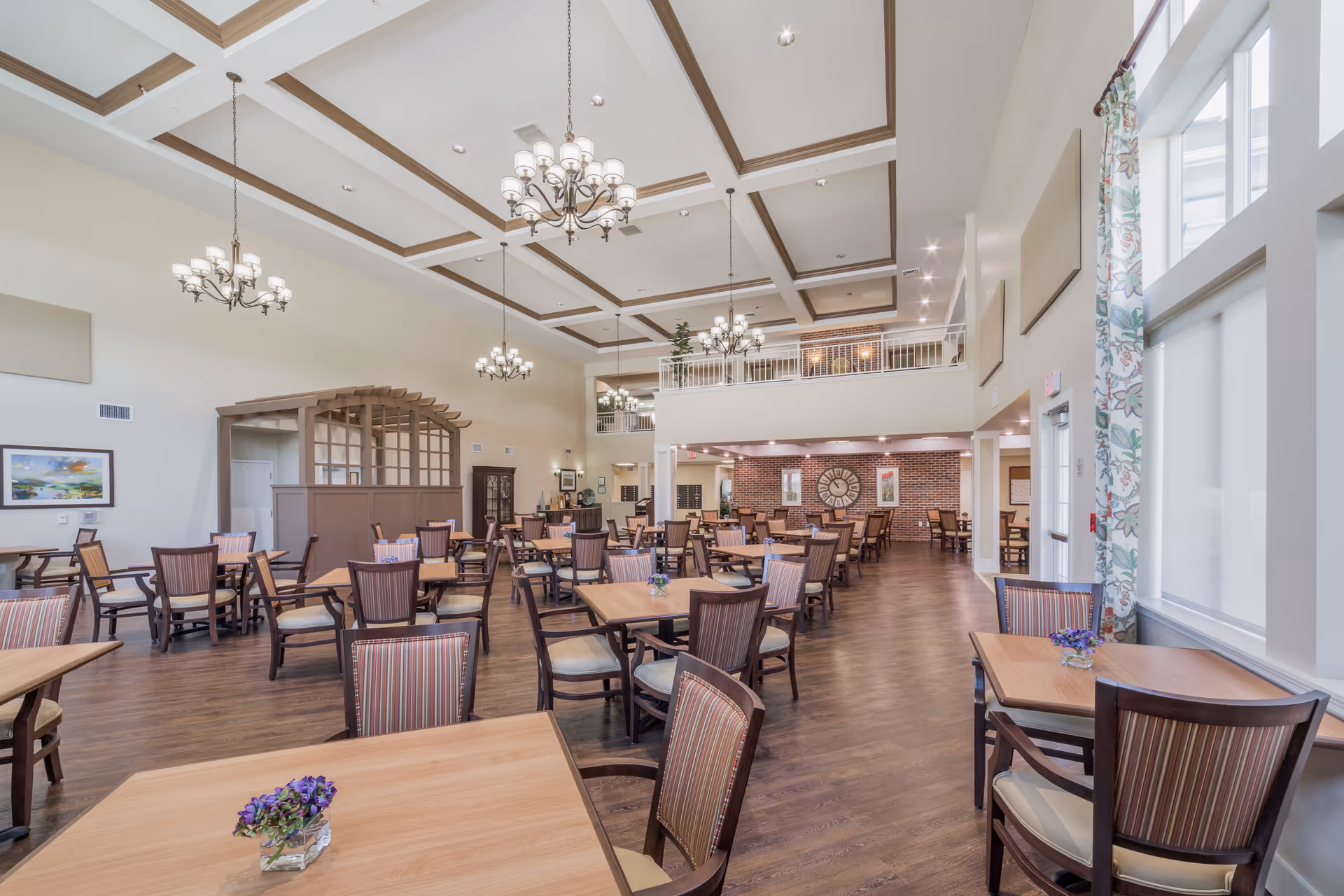 A spacious dining room with multiple wooden tables and chairs arranged neatly. The room features high ceilings with decorative beams and several chandeliers hanging down. Large windows with floral curtains allow natural light to fill the space. The floor is wooden, and there are small flower arrangements on some tables. A balcony area overlooks the dining space, and a large clock is mounted on a brick accent wall in the background.
