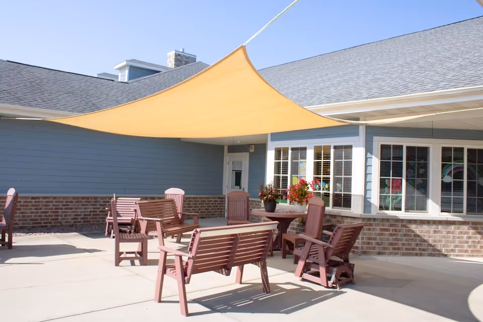 Outdoor patio area with wooden benches and chairs arranged around a round table with potted flowers. A large beige shade sail is stretched overhead, attached to the building. The building has blue siding with brick accents and several windows.