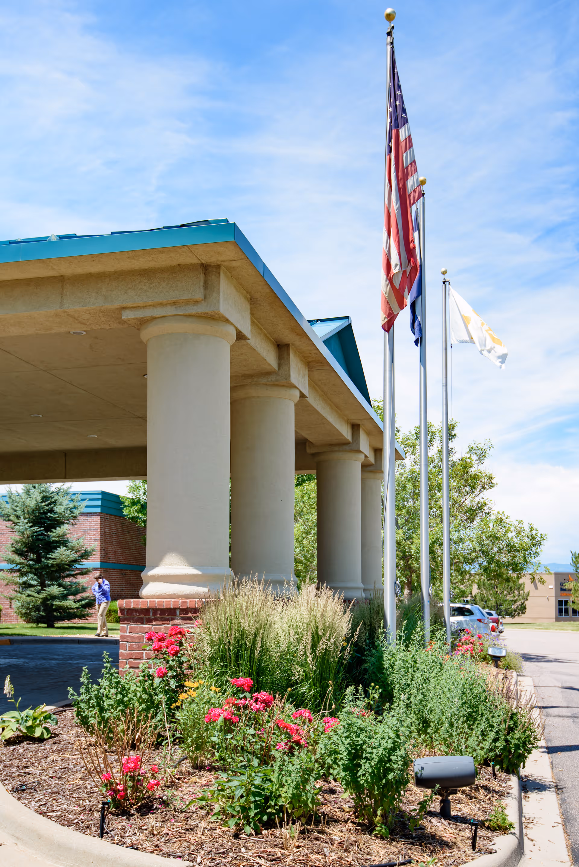 Covered entrance with large columns, flagpoles flying flags, and landscaped flower beds in front of the facility.