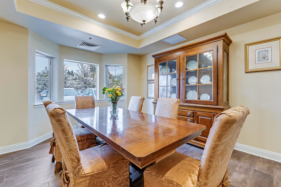 Sunlit dining room with a wooden table, upholstered chairs, a china cabinet, and a vase of flowers on the table.