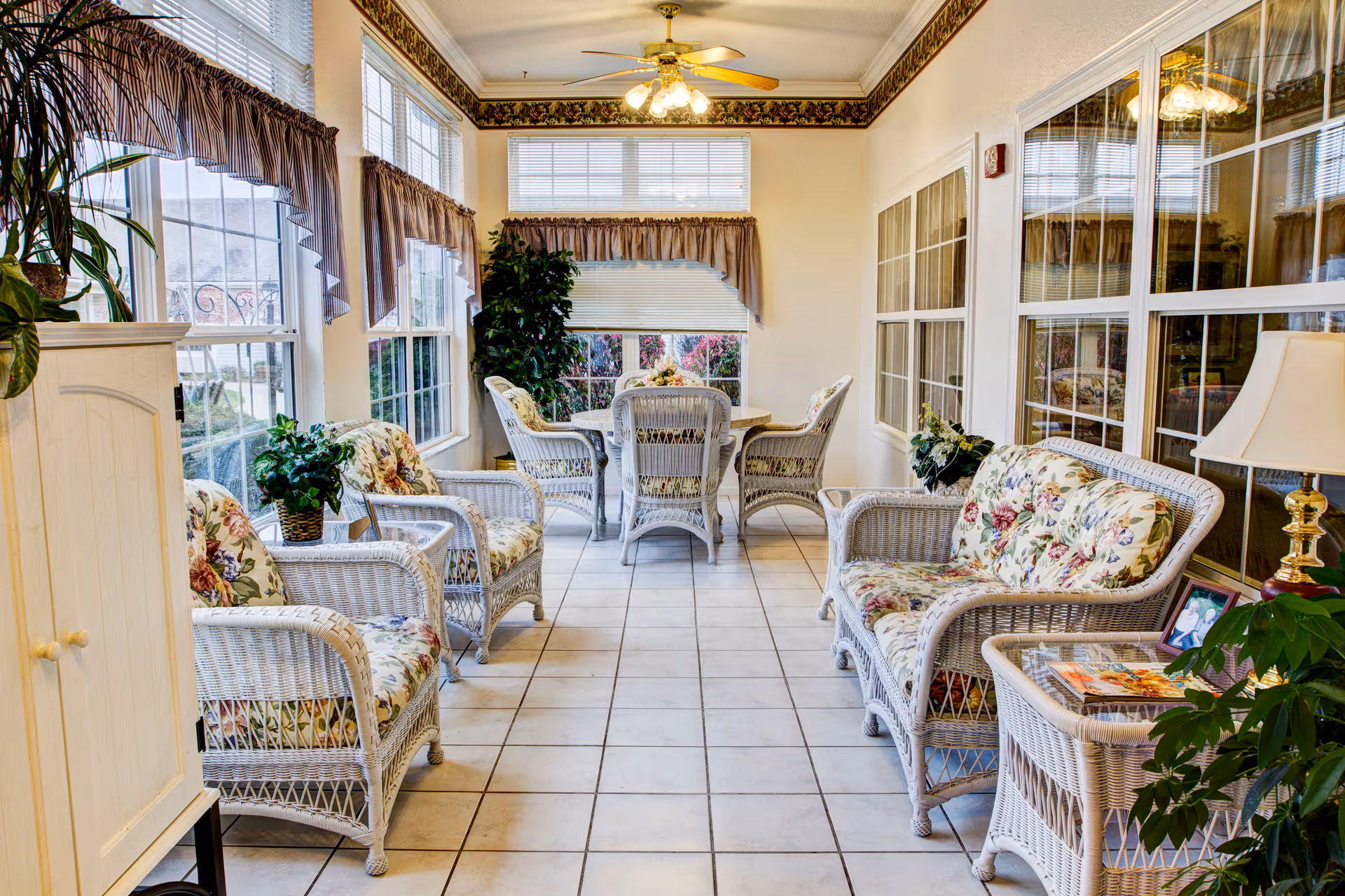 Sunlit sitting room with white wicker chairs and a small round dining table, floral cushions, and large windows.