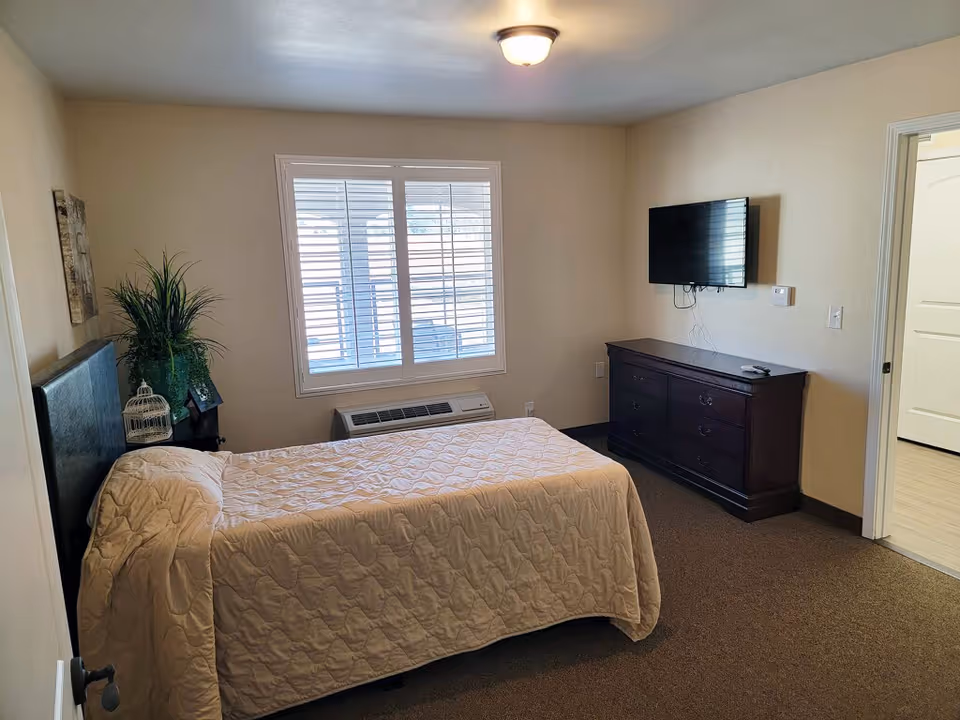 A bedroom with a bed covered in a beige quilt, a dark wooden dresser with a flat-screen TV mounted above it, a window with white blinds, a small nightstand with a plant and decorative items, and a door leading to another room.