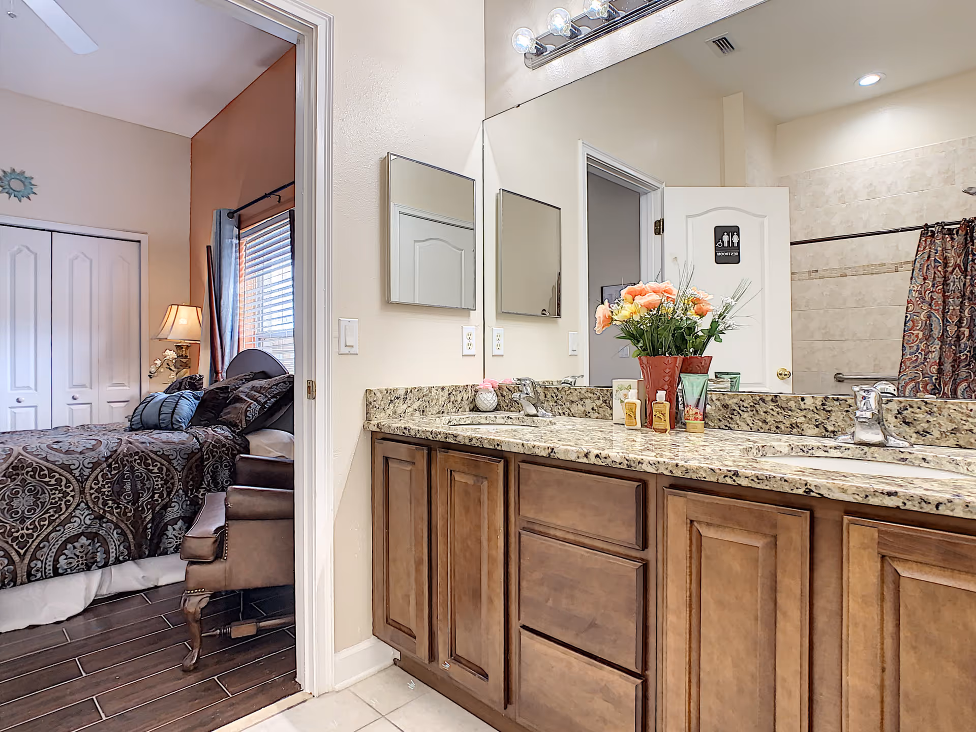 View of a bathroom with a granite countertop, wooden cabinets, two sinks, and a large mirror above. A vase with flowers and toiletries are placed on the counter. The bathroom door is open, showing a shower area with a patterned curtain and a sign on the door. To the left, there is a doorway leading to a bedroom with a bed, pillows, a lamp, and a window with blinds.