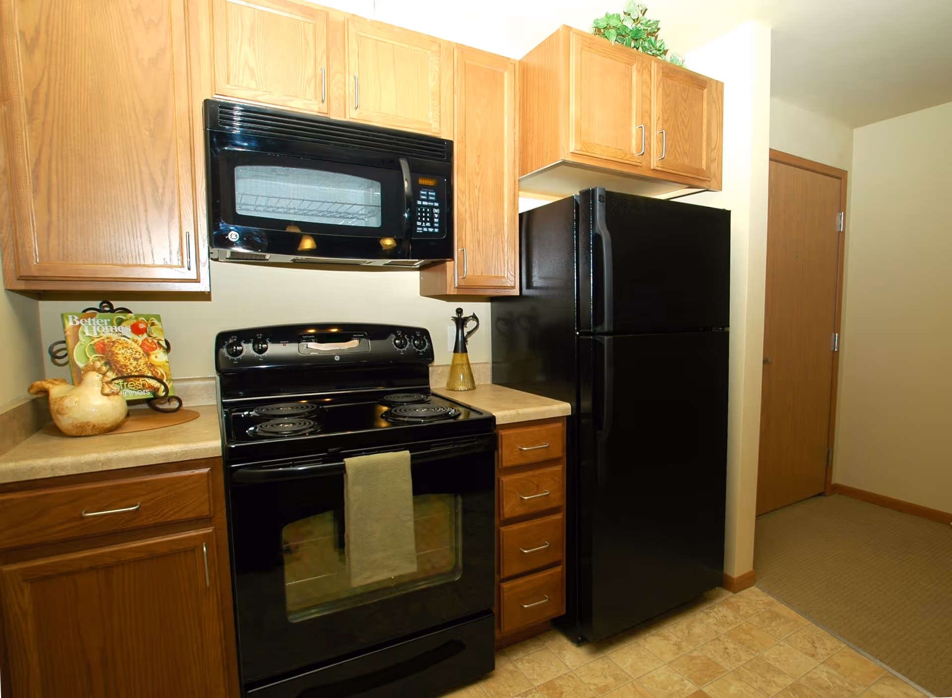 A kitchen area featuring wooden cabinets, a black microwave mounted above a black electric stove with a towel hanging on the oven door, and a black refrigerator. The countertop has a decorative ceramic chicken and a cookbook stand with a cookbook. There is a doorway leading to another room in the background.