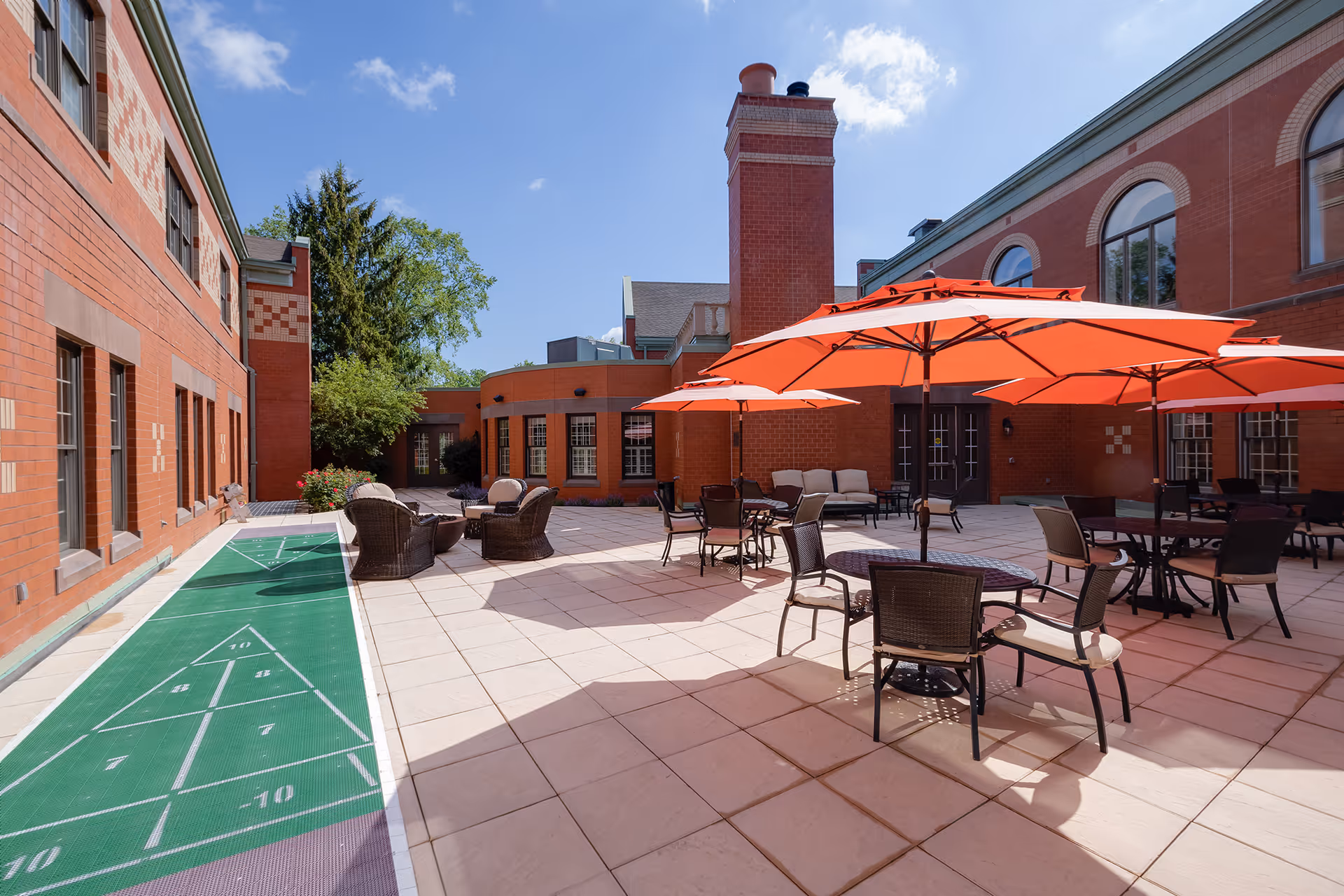 Outdoor courtyard area at Evergreen Woods featuring several tables with orange umbrellas, chairs, and a shuffleboard court along the left side. The courtyard is surrounded by red brick buildings under a clear blue sky.