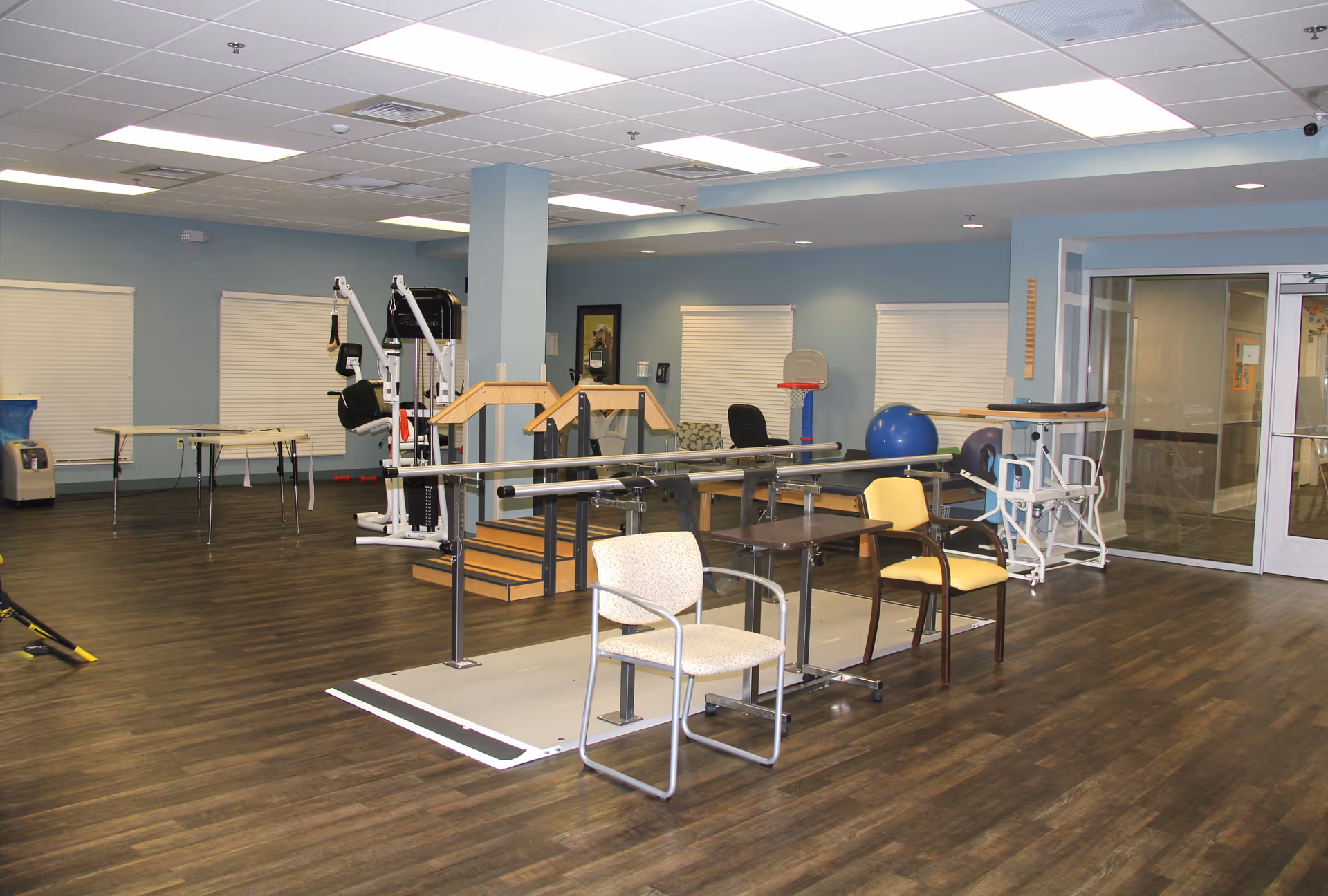 A spacious rehabilitation or physical therapy room with exercise equipment, parallel bars for walking practice, chairs, and tables. The room has blue walls, wood flooring, and a drop ceiling with fluorescent lighting. There are windows with closed blinds and a glass door leading to another area.
