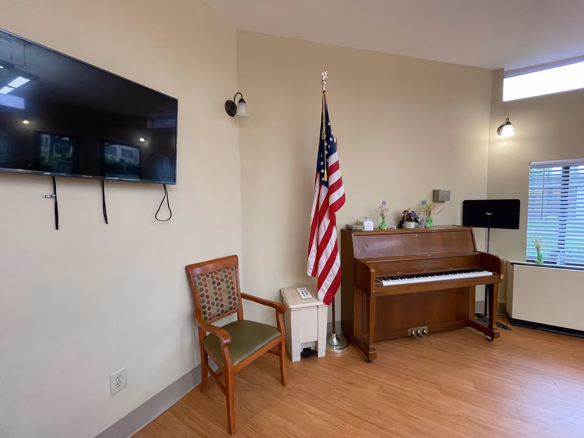 A corner of a room with a wooden upright piano, an American flag on a stand, a wooden chair with a patterned cushion, a wall-mounted flat screen TV, and a window with blinds. The floor is wood, and there are two wall-mounted light fixtures.