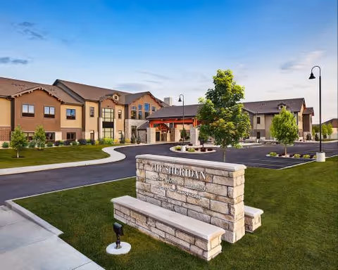 Exterior view of a two-story assisted living facility building with a well-maintained lawn, trees, and a paved driveway. A stone sign in the foreground reads 'The Sheridan, Assisted Living & Memory Care'. The sky is clear with a few clouds.