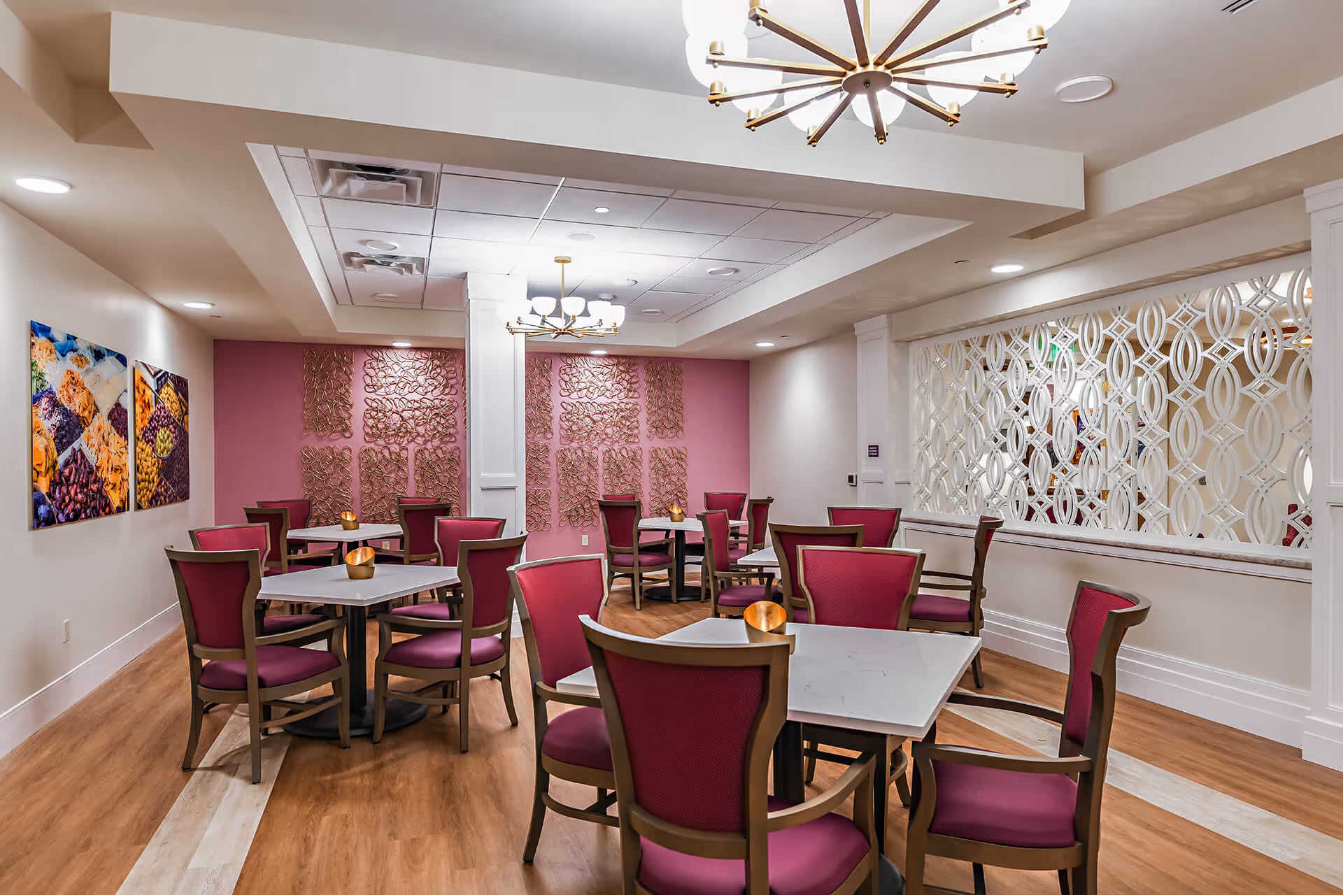 Dining room with several square tables surrounded by magenta-upholstered chairs, chandeliers, and decorative wall panels.