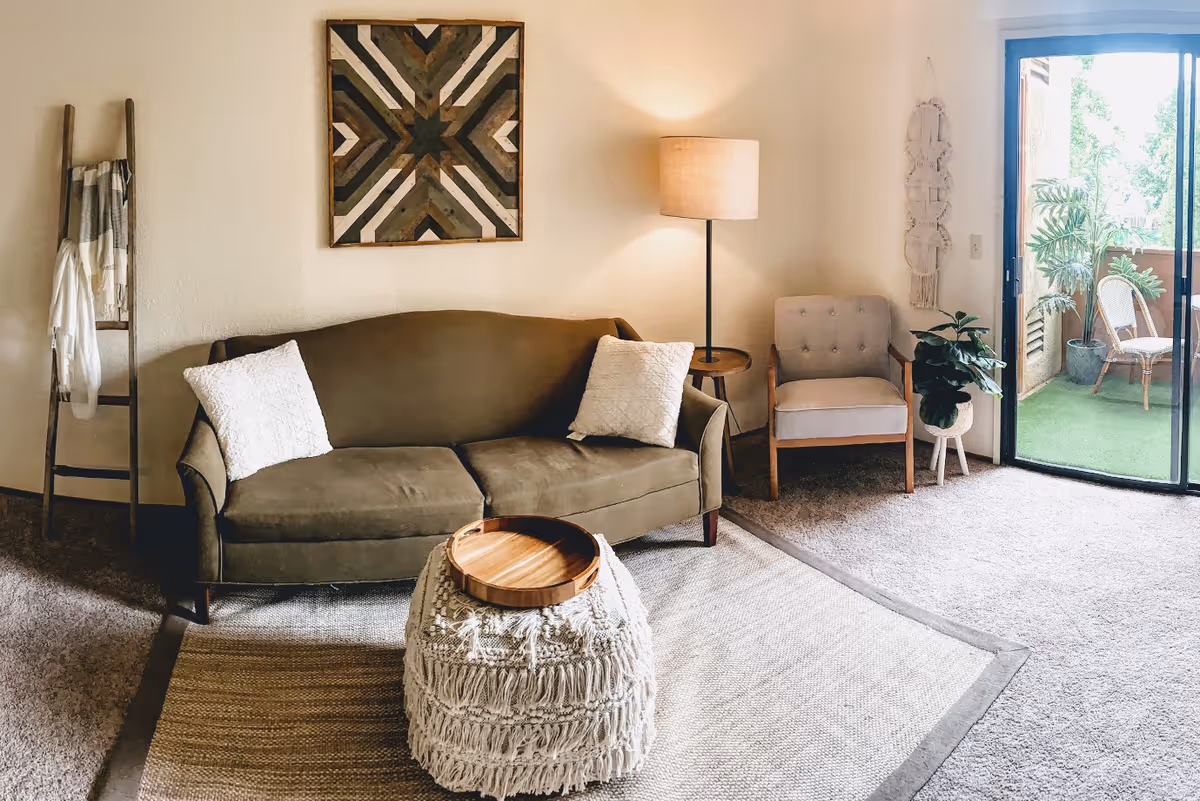 A cozy living room with a green sofa adorned with two white pillows, a textured ottoman with a wooden tray on top, a wooden ladder with blankets hanging on it, a floor lamp beside a beige armchair, a potted plant, and a sliding glass door leading to a balcony with outdoor chairs and plants.