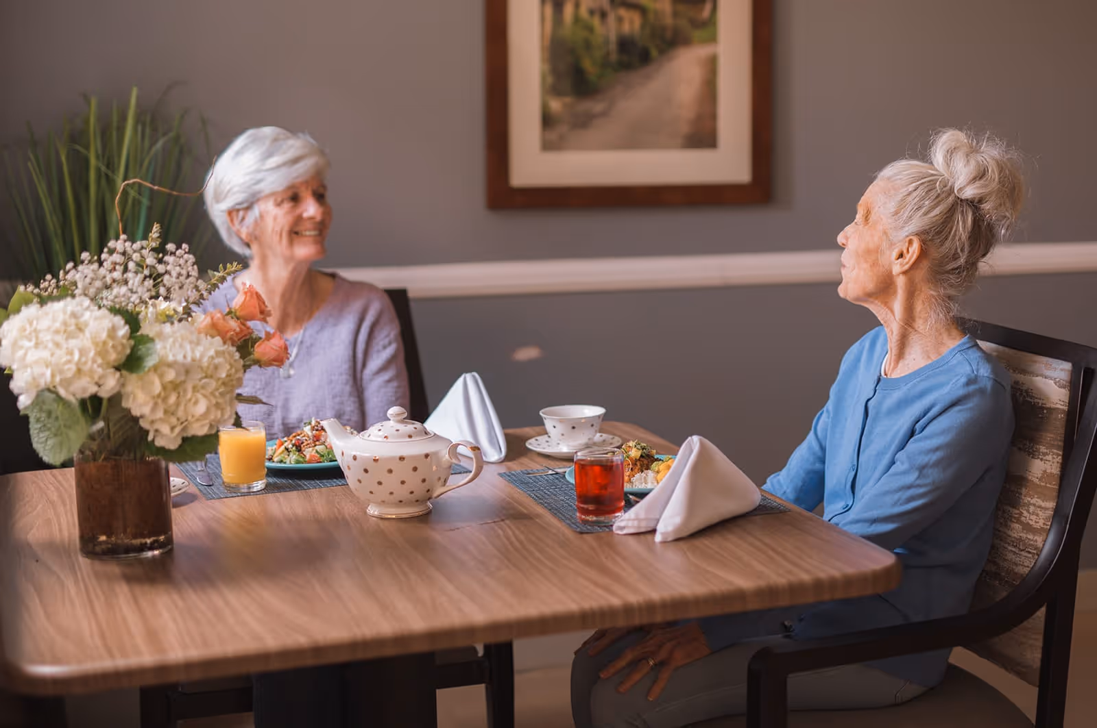 Two elderly women sitting at a wooden dining table in a cozy room, enjoying a meal together. The table is set with a teapot, cups, glasses of juice, plates of food, and folded white napkins. A vase with white and pink flowers is also on the table. A framed picture hangs on the gray wall behind them.