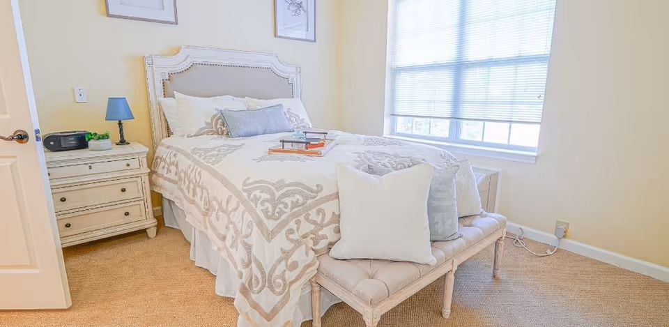 Bright, softly decorated bedroom with a patterned bed, upholstered bench at the foot, nightstand, and a large window.