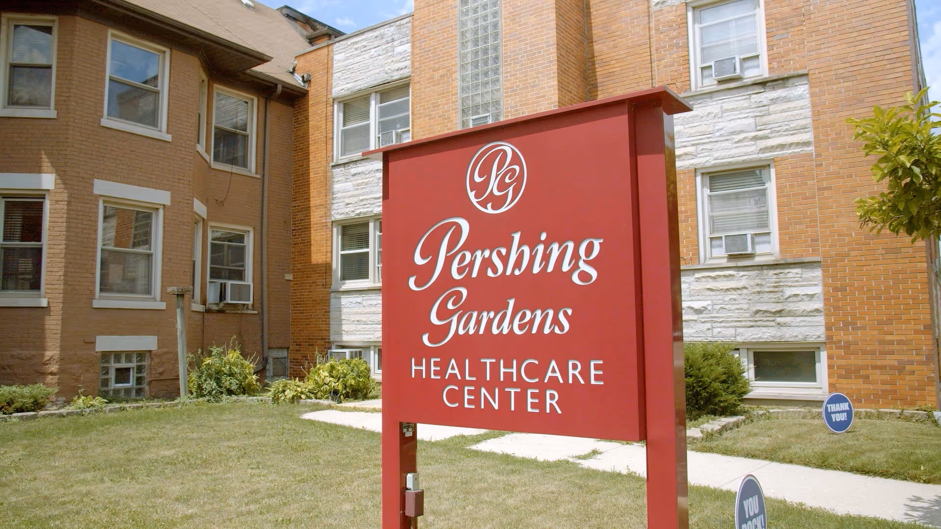 A red sign in front of a brick building reads 'Pershing Gardens Healthcare Center'. The building has multiple windows with air conditioning units and is surrounded by a grassy area with a sidewalk and some small bushes.