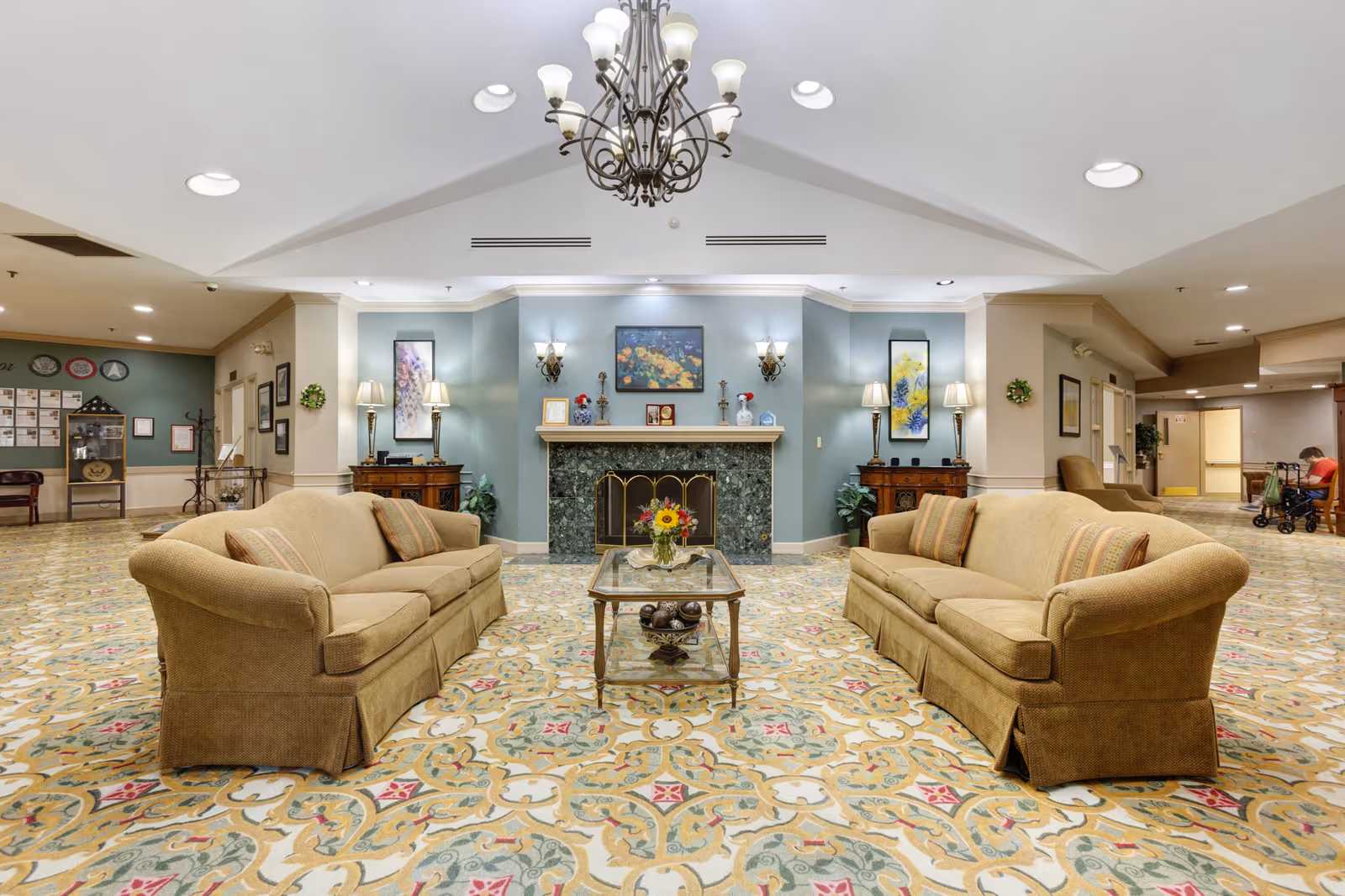 A spacious and well-lit living room area in Independence Village of Plymouth featuring two beige sofas facing each other with a glass coffee table in between. The room has a decorative patterned carpet, a green marble fireplace with framed artwork above it, and two wooden side tables with lamps and plants. A chandelier hangs from the ceiling, and there is a person sitting in the background near a walker.
