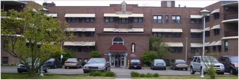Brick senior living facility with a central entrance and cars parked in front.