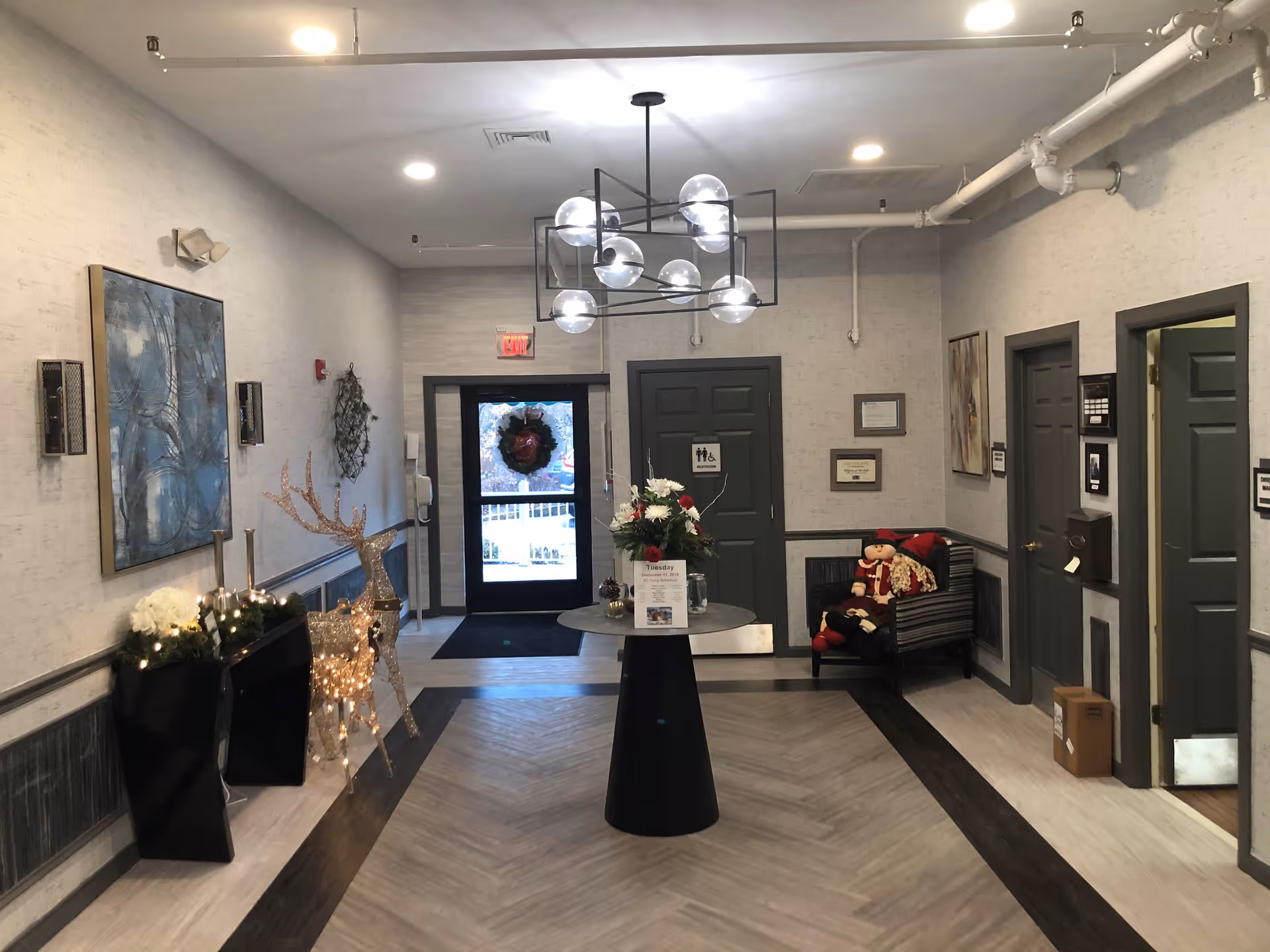 Interior view of a hallway or lobby area in a senior living facility with a modern chandelier, a round table with a flower arrangement, holiday decorations including a lighted reindeer figure, a wreath on the door, and a chair with festive dolls. Several doors and framed pictures are visible along the walls.