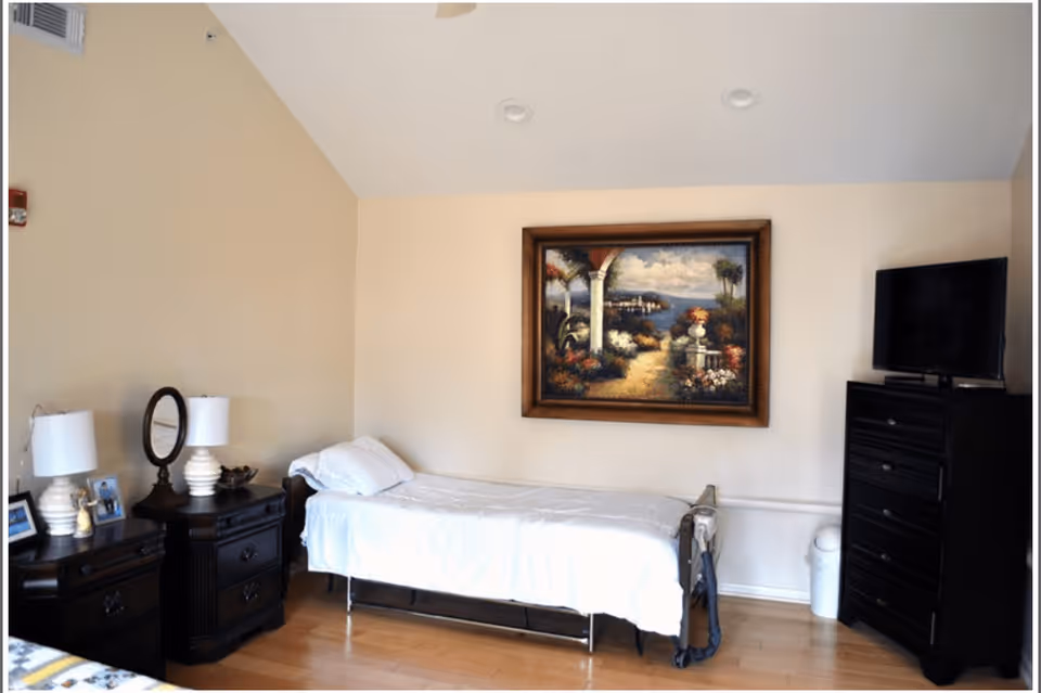 A neatly arranged bedroom in an assisted living facility featuring a single bed with white bedding, two dark wooden nightstands with lamps and framed photos, a dark wooden dresser with a flat-screen TV on top, and a large framed painting of a scenic garden and ocean view on the wall.