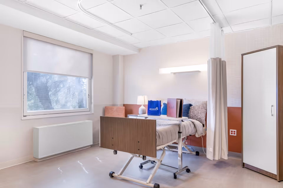 A bright and clean patient room in a healthcare facility featuring a hospital bed with a wooden headboard and footboard, a bedside table with a lamp and some items on it, a window with a roller shade partially down, a privacy curtain on a ceiling track, a wardrobe with a white door, and a cushioned chair in the corner.