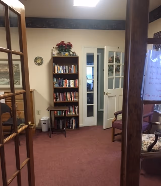 A small interior common room with a tall bookshelf, glass-paneled doors, chairs, and maroon carpet.
