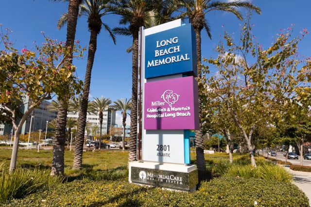 Outdoor view of a tall signpost with three panels reading Long Beach Memorial, Miller Children's & Women's Hospital Long Beach, and 2801 Atlantic, surrounded by palm trees and greenery under a clear blue sky.