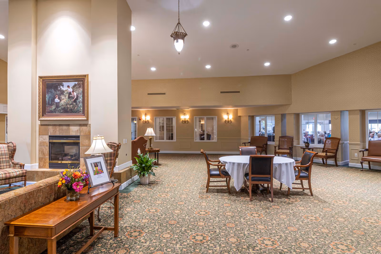 A spacious, well-lit common area in a senior living facility with patterned carpet, a round table covered with a white tablecloth surrounded by chairs, several armchairs along the walls, a wooden table with a lamp, flowers, and a framed photo, and a fireplace with a painting above it.