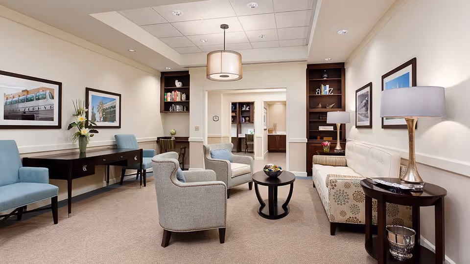 A well-lit living room area with beige walls and carpet, featuring a patterned sofa, two gray armchairs, a round black coffee table, a side table with a lamp, and a desk with two blue chairs. The room has framed pictures on the walls, built-in dark wood shelves, and a modern ceiling light fixture.