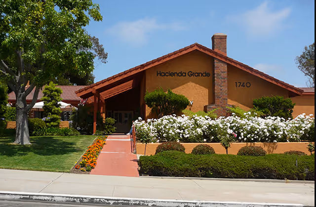 Front exterior view of Hacienda Grande Senior Assisted Living building with a brown stucco facade, a red-tiled roof, a brick chimney, and the address number 1740. The entrance is surrounded by well-maintained landscaping including green bushes, white flowers, orange flowers along the walkway, and trees under a clear blue sky.