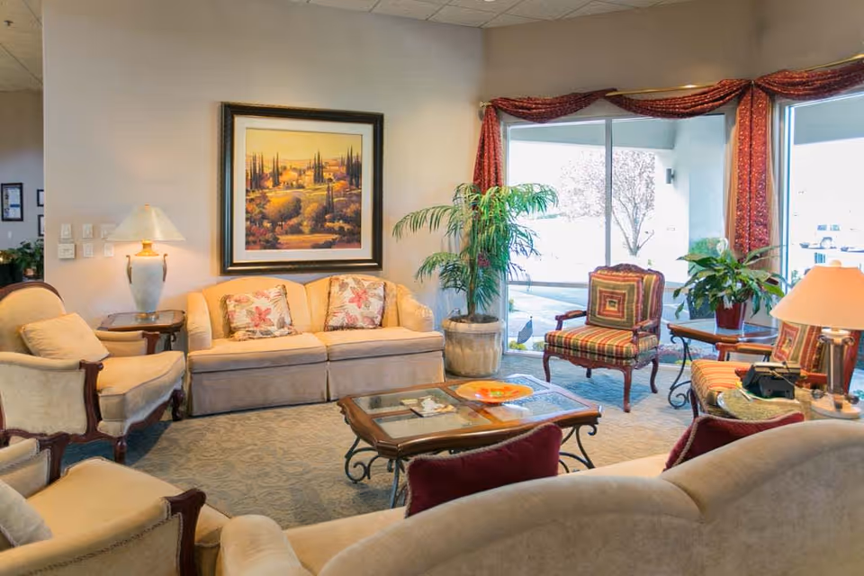 A cozy living room with beige sofas and armchairs arranged around a glass coffee table. The room features a large framed landscape painting on the wall, potted plants, and large windows with red curtains letting in natural light.