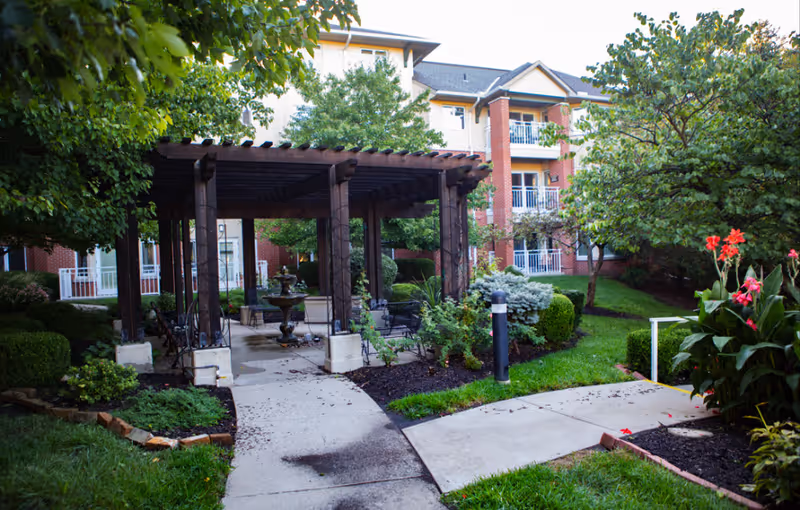 Outdoor garden area at Claridge Court featuring a wooden pergola with seating underneath, surrounded by well-maintained greenery, bushes, trees, and colorful flowers. A multi-story brick and beige building with balconies is visible in the background.
