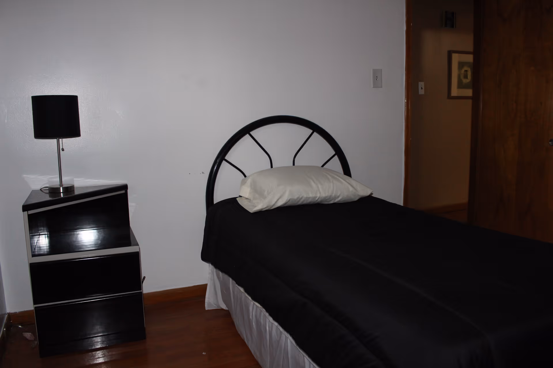 A simple bedroom with a single bed featuring a black metal headboard, a white pillow, and a black bedspread. Next to the bed is a black nightstand with two drawers and a black lamp on top. The room has wooden flooring, white walls, and a wooden door partially open showing a hallway with a framed picture on the wall.