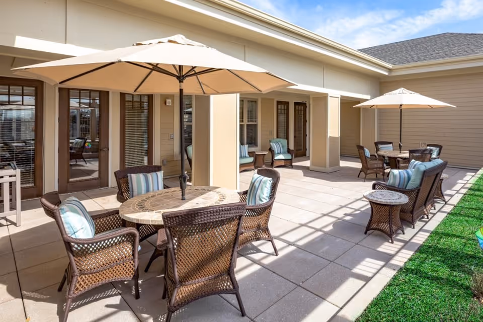 Outdoor covered patio with wicker tables, chairs, umbrellas and cushioned seating outside a senior living facility.