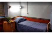 A small bedroom with a single bed covered in a blue quilted bedspread. Next to the bed is a wooden nightstand with a potted plant on top. A wall-mounted light fixture is above the bed, and a window with closed blinds is partially visible on the left side.