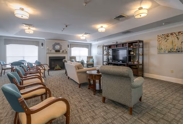 A spacious living room area with multiple upholstered chairs and sofas arranged around a wooden coffee table and a fireplace. There is a large TV on a dark wooden entertainment unit with shelves holding decorative items and books. The room has carpeted flooring, soft lighting from ceiling fixtures, and two large windows with blinds allowing natural light to enter. The walls are decorated with framed artwork.