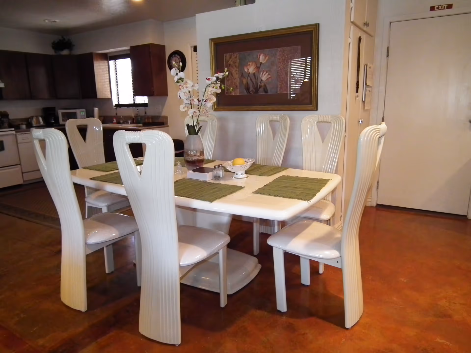 Open dining area showing a white table set with green placemats and six white high-backed chairs, with a kitchen in the background.