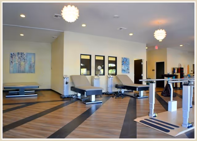 A spacious wellness or therapy room with wooden flooring and modern treatment tables. The room features light yellow walls adorned with blue-themed artwork and three rectangular mirrors. There are medical or therapy devices near the tables and parallel bars for physical therapy exercises. The ceiling has recessed lighting and two decorative pendant lights.