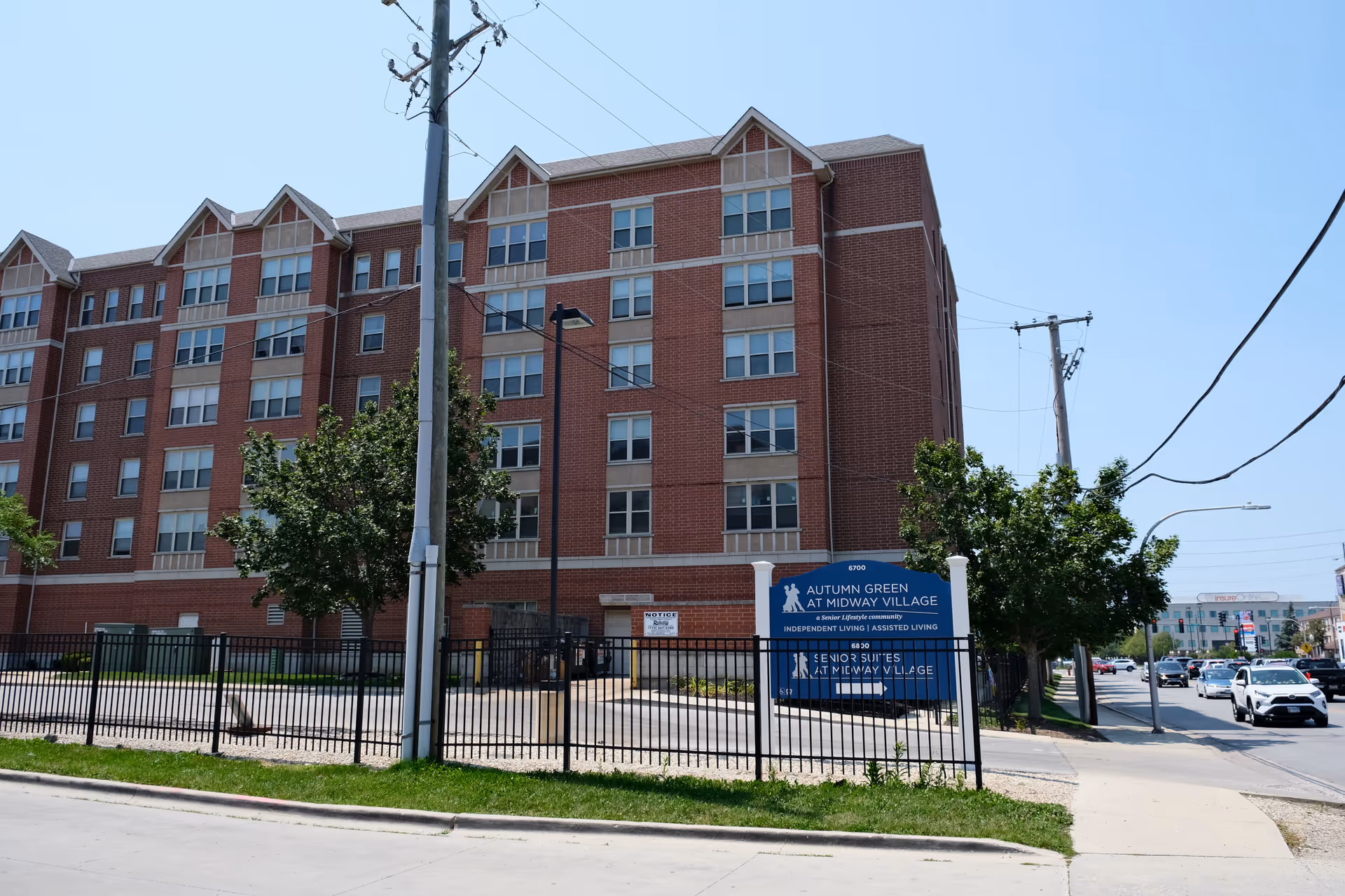 Exterior view of a multi-story brick building with multiple windows, surrounded by a black metal fence and some trees. A blue sign in front reads 'Autumn Green at Midway Village, a Senior Lifestyle community, Independent Living | Assisted Living'. Cars are visible on the street to the right under a clear blue sky.