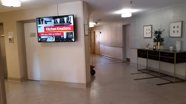 Interior hallway/lobby with a wall-mounted TV showing 'Kitchen Kreations', a console table with decorations on the right, and a corridor leading to rooms.
