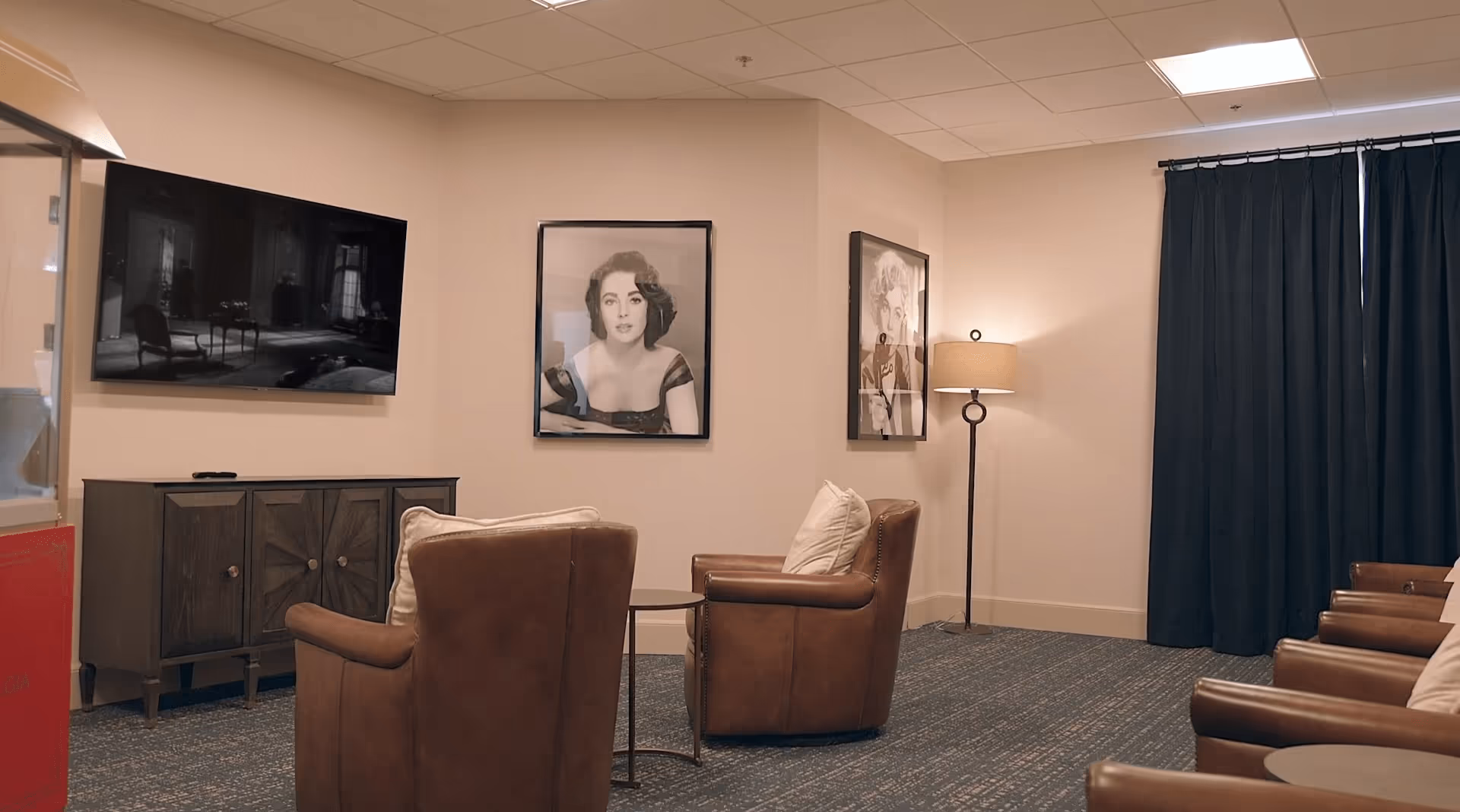 A cozy lounge with leather armchairs, a wall-mounted TV, framed black-and-white portraits, a floor lamp and dark curtains.