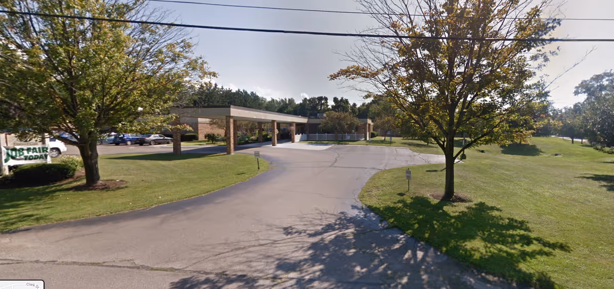 Exterior view of Absolut Care of Westfield facility showing a driveway leading to a covered entrance. The building is single-story with brick walls. There are trees and green lawns on both sides of the driveway, and a sign on the left side advertising a job fair today.