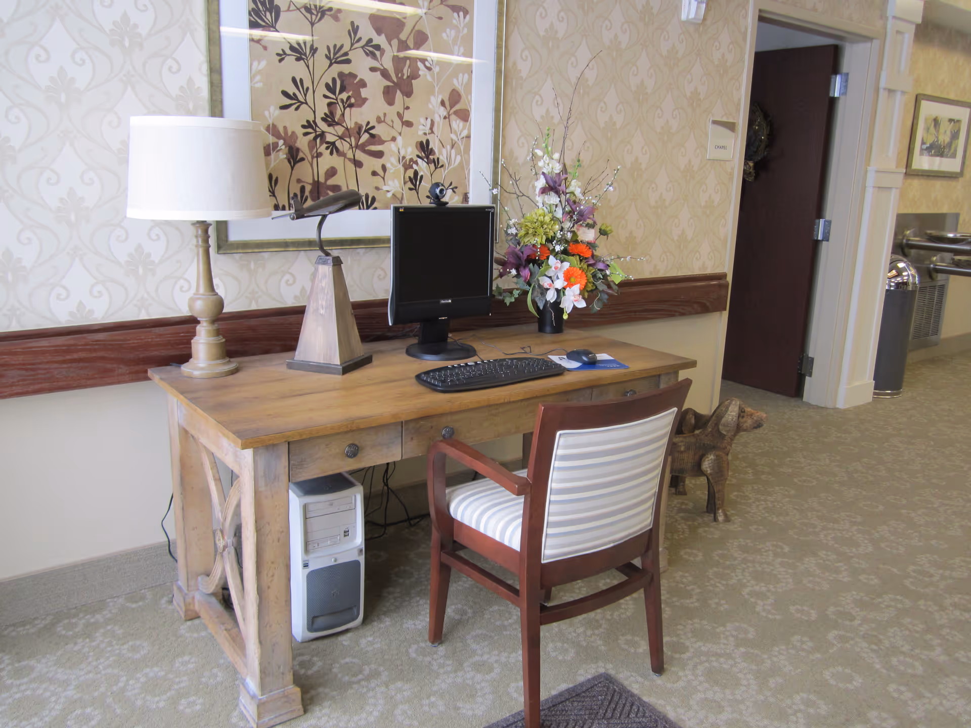 A wooden desk with a computer monitor, keyboard, mouse, lamp, and a vase of colorful flowers on top. A wooden chair with a striped cushion is placed in front of the desk. The desk is set against a wall with patterned wallpaper and a framed floral artwork. The floor is carpeted with a floral pattern, and there is a hallway visible to the right with a door and some framed pictures on the wall.